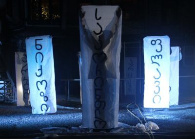Zdjęcie przedstawia białe płachty z gruzińskimi napisami nazw wsi i miejscowości. Są trzymane przez grupę aktorów regularnie rozstawionych na nocnym, oświetlonym niebieskim światłem, placu. To scena ze spektaklu Dom Granica.