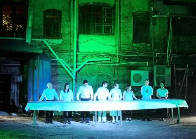 Zdjęcie przedstawia grupę kobiet i mężczyzn w białych kostiumach. Stoją przy długim stole, zasłanym białym obrusem. Są na oświetlonym reflektorami na niebiesko i zielono dziedzińcu przemysłowych budynków. Jest noc. Z tyłu wentylatory i liczne rynny.