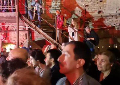 Zdjęcie przedstawia tłum publiczności podczas spektaklu Dom Granica fundacji Art Dżankszon w Tbilisi. Stoją pośród poprzemysłowych budynków i na przylegających do nich metalowych schodach. Są oświetleni czerwonym światłem z reflektorów.