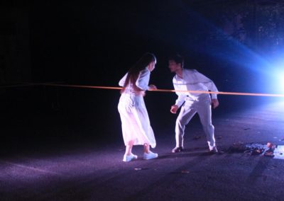Zdjęcie przedstawia parę w białych kostiumach, tańczącą gruziński taniec. Pochylają się ku sobie stojąc po dwóch stronach rozciągniętej, czerwonej linii. Są na oświetlonym reflektorami na niebiesko placu. Jest noc.