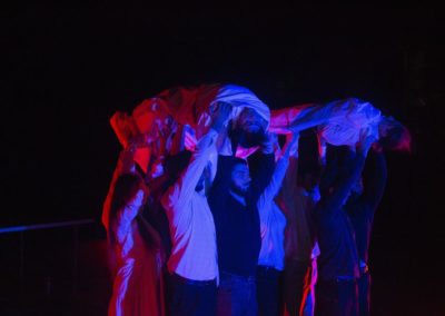Zdjęcie przedstawia grupę ludzi unoszącą na wzniesionych rękach ciała dwóch ofiar - aktorów wcielających się w tę rolę. Są oświetleni reflektorami na różowo i niebiesko. Jest noc. To fragment spektaklu Dom Granica Fundacji Art Dżankszon.