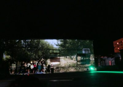 Zdjęcie przedstawia scenę ze spektaklu Dom Granica. Na budynku wyświetlany jest film. Wokół grupa widzów. Jest noc.
