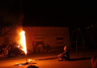 Zdjęcie przedstawia mężczyznę siedzącego na ziemi przed wielką płonącą pochodnią postawioną na placu pomiędzy przemysłowymi budynkami. To scena ze spektaklu Dom Granica. Wokół grupa widzów. Jest noc.