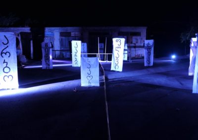Zdjęcie przedstawia białe płachty z gruzińskimi napisami nazw wsi i miejscowości. Są trzymane przez grupę aktorów regularnie rozstawionych na nocnym, oświetlonym niebieskim światłem, placu pomiędzy przemysłowymi budynkami. To scena ze spektaklu Dom Granica.