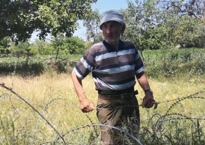 Zdjęcie przedstawia starszego pana stojącego w ogrodzie z sadem za drutem kolczastym. Jest w czapeczce i pasiastej bluzce. Rozmawia z osobą go fotografującą.