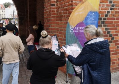 Zdjęcie przedstawia przemierzających przez Olsztyn uczestników spaceru sąsiedzkiego Fundacji Art Dżankszon. Przechodzą przez arkady starej, ceglanej bramy wysokiej. Przy niej wielokolorowy banner z napisem Sąsiedztwo moje miejsce na mapie.