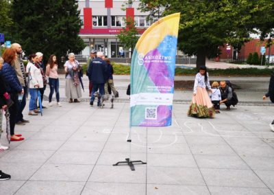 Zdjęcie przedstawia grupę kilkunastu osób z różnych pokoleń na miejskim placu. Wśród nich romskie dziewczynki w tradycyjnych barwnych sukniach. Na środku placu wielokolorowy banner z napisem Sąsiedztwo moje miejsce na mapie.