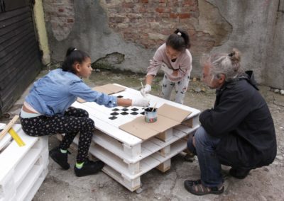 Zdjęcie przedstawia dwie romskie dziewczynki malujące szachownicę na stole z palet. Ich pracę obserwuje mężczyzna o srebrnych włosach spiętych w koczek. Znajdują się na podwórku, przy odrapanej do cegieł ścianie.