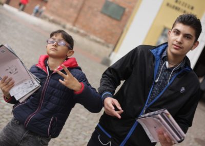 Zdjęcie przedstawia dwóch ciemnowłosych chłopców, trzymających w dłoniach foldery z mapami projektu Sąsiedztwo. Są w sportowych kurteczkach. Po lewej stronie młodszy, w okularach, z wykonywanym lewą dłonią gestem wiktorii, po prawej starszy. Wzrok mają skierowany wprost w obiektyw.