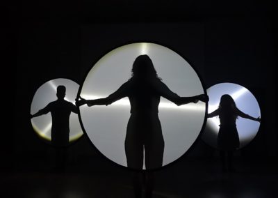 Zdjęcie przedstawia cienie dwóch kobiet i mężczyzny na tle plansz przypominających płyty kompaktowe znajdujących się na czarnym tle. Każda z postaci ma symetrycznie rozłożone ręce.