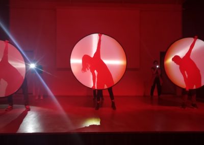 Zdjęcie przedstawia trzy postacie stojące na scenie zasłonięte okrągłymi blendami. Widzimy tylko ich cienie. Są pochyleni w lewą stronę, ręce mają rozłożone w górę i dół. Dominują kolory czerwieni i różu, scena jest oświetlona przyciemnionymi reflektorami.