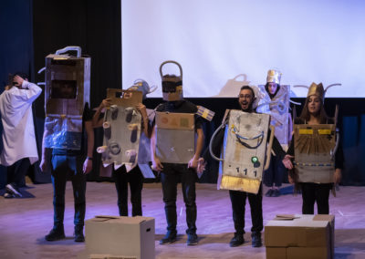 Zdjęcie przedstawia grupę młodych aktorów i aktorek w kartonowych kostiumach robotów stojących na teatralnej scenie.