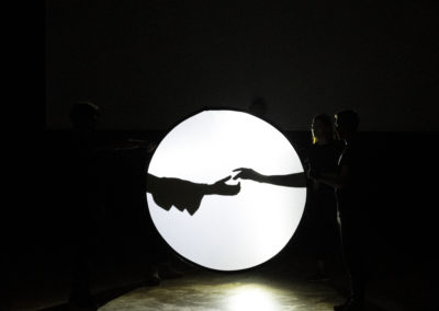 Zdjęcie przedstawia scenę teatru cieni. Na białej blendzie przypominającej księżyc, czarne odbicie dwóch usiłujących dosięgnąć siebie dłoni. Ta po lewej ma rękaw z falbanami, ta po prawej jest drobniutka. Tablica trzymana jest przez trzy osoby stojące w cieniu.