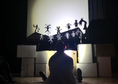 Zdjęcie przedstawia siedzącego do nas tyłem, na podłodze, mężczyznę w kitlu. Wpatruje się w ekran na którym trwa pokaz teatru cieni. Kukiełki są wycięte z papieru roboty i potwory. Przed nim stoją białe pudła, a obok, w cieniu siedzi inna osoba.