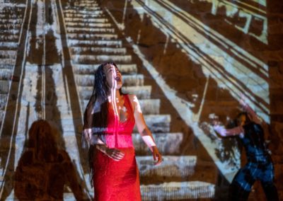 Zdjęcie przedstawia scenę ze spektaklu. Na pierwszym planie śpiewa atrakcyjna brunetka w czerwonej sukni z dekoltem. Ma rozłożone dłonie. Za nią na ścianie wyświetlana jest animacja przedstawiająca ruchome schody. W prawym narożniku stoi odwrócona tyłem, skulona postać opierająca się o ścianę.