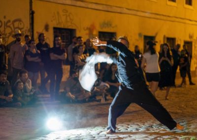 Zdjęcie przedstawia dynamiczny ruch mężczyzny trzymającego w dłoni białą szmatę. Jest rozmazana. Stoi boso w rozkroku na brukowanej ulicy, zamachuje się rękoma. Jest ciemno. Wokół są zgromadzeni widzowie, za nimi wymalowany graffiti budynek.
