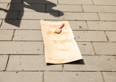 Zdjęcie przedstawia tekturę leżącą na chodniku, na której pędzlem przy użyciu wody wykonano napis, zanikający pod wpływem słońca. Trudno już odczytać napisane słowo.