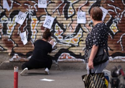 Zdjęcie przedstawia akcję uliczną w której dziewczyna przykleja do muru zamalowanego przez graffiti kartki z głoskami en oraz eu. Kuca przy ścianie. Na pierwszym planie zwrócona w jej stronę starsza kobieta z torbami zakupowymi.