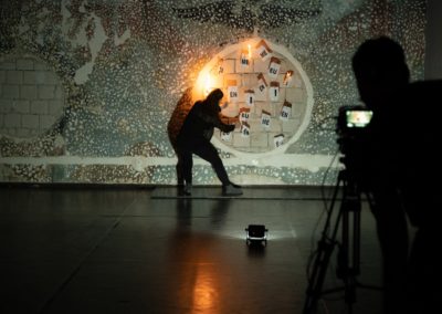 Zdjęcie przedstawia moment nagrywania spektaklu zatytułowanego jesteśmy gdzie. Dziewczyna podpala przyklejone do wymalowanego na ścianie okręgu kartki z głoskami. W prawym rogu znajduje się zarys kamerzysty i kamera na statywie.
