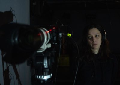 Zdjęcie przedstawia operatorkę filmową stojącą za kamerą skierowaną w stronę obiektywu aparatu. Młoda kobieta ma na uszach duże słuchawki, ciemne rozpuszczone włosy. Siedzi spoglądając na kamerę w ciemnym pomieszczeniu.