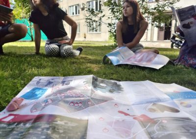 Na zdjęciu dwie nastolatki siedzą na trawie przysłuchując się wypowiedzi osoby spoza kadru. Na pierwszym planie znajduje się rozłożona mapa sąsiedzka Nowej Huty z zaznaczonymi znaczącymi punktami i umieszczonymi fotografiami dzielnicy. W tle znajduje się budynek.