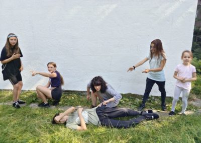 Zdjęcie przedstawia szóstkę dzieci w wieku od kilku do kilkunastu lat na trawie przy białej, przygotowanej do powstania muralu ścianie. Są w ruchu, zadowolone. Dziewczynki wskazują na siebie palcami, chłopiec leży na trawie zasłaniając dłonią twarz.