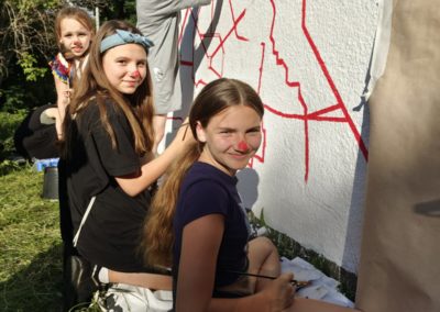 Zdjęcie przedstawia trzy dziewczynki z wymalowanymi na czerwono noskami. Siedzą przy ścianie na której powstaje mural. Czerwone linie składające się na zarys mapy. Dziewczynki są zwrócone w stronę aparatu, uśmiechnięte, w dłoniach trzymają pędzle. Za nimi widać dwie kobiety malujące ścianę.
