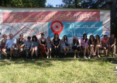 Zdjęcie przedstawia grupę kilkunastu osób w różnym wieku siedzących na krzesłach na tle muralu w kolorach różowy, niebieski, czerwony i biały z napisami Sąsiedztwo, Nowa Huta.