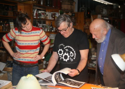 Zdjęcie przedstawia trzy osoby z zaciekawieniem przyglądające się książce z archiwalnymi fotografiami. Kobieta i dwóch panów znajdują się w pomieszczeniu wypełnionym pamiątkami.